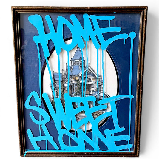 A framed art print featuring a blue and white Victorian house behind bright blue dripping graffiti-style text that reads 'HOME SWEET HOME' on a dark blue background.